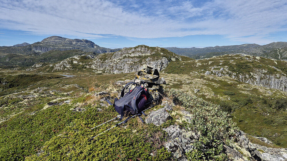 Sekken ligger inntil toppvarden på Døvlingstjørnfjellet Sørøst (1117). Nærmeste topp bak, rett over toppvarden og sekken, er høyeste Døvlingstjørnfjellet (1130). Lengre bak til venstre ruver Vindeggen (1516).